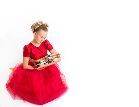 Caucasian girl with blond curls wearing a red party dress sits on the floor holding a nutcracker doll in her arms on an isolated white background