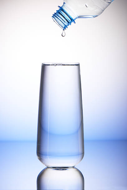 Drop of water falling from plastic bottle into full drinking glass with reflection on white and blue background