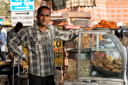 Jaipur, India - December 8, 2015: A young man standing at his street stand located just on the side of a busy street. where he prepares and sells food, Jaipur, Rajasthan, India. Jaipur is the capital and largest city of the Indian state of Rajasthan in Northern India.