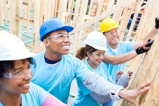 Diverse friends are volunteers working together to build home for charity. They are confidently putting up a wall in the home. They are all holding the wall. They are wearing hard hats, safety glasses and light blue volunteer t-shirts.