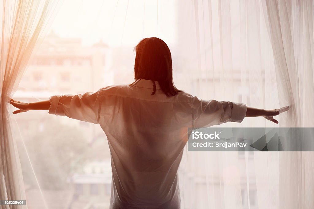 It's A Brand New Day! Rear view of a woman waking up, standing by the bedroom window and opening the curtains. New Stock Photo