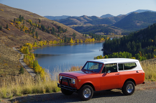 A bright orange car parked above a blue lake on a fall day. Leaves are turning in the background.