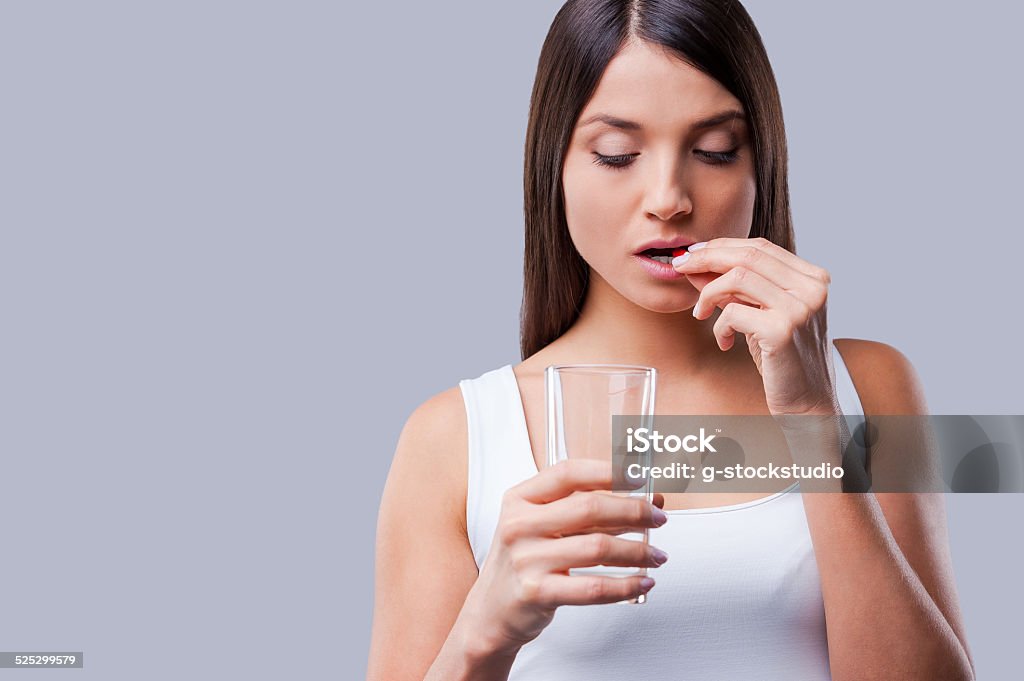 Woman taking pills. Depressed young woman taking a pill while standing against grey background Women Stock Photo