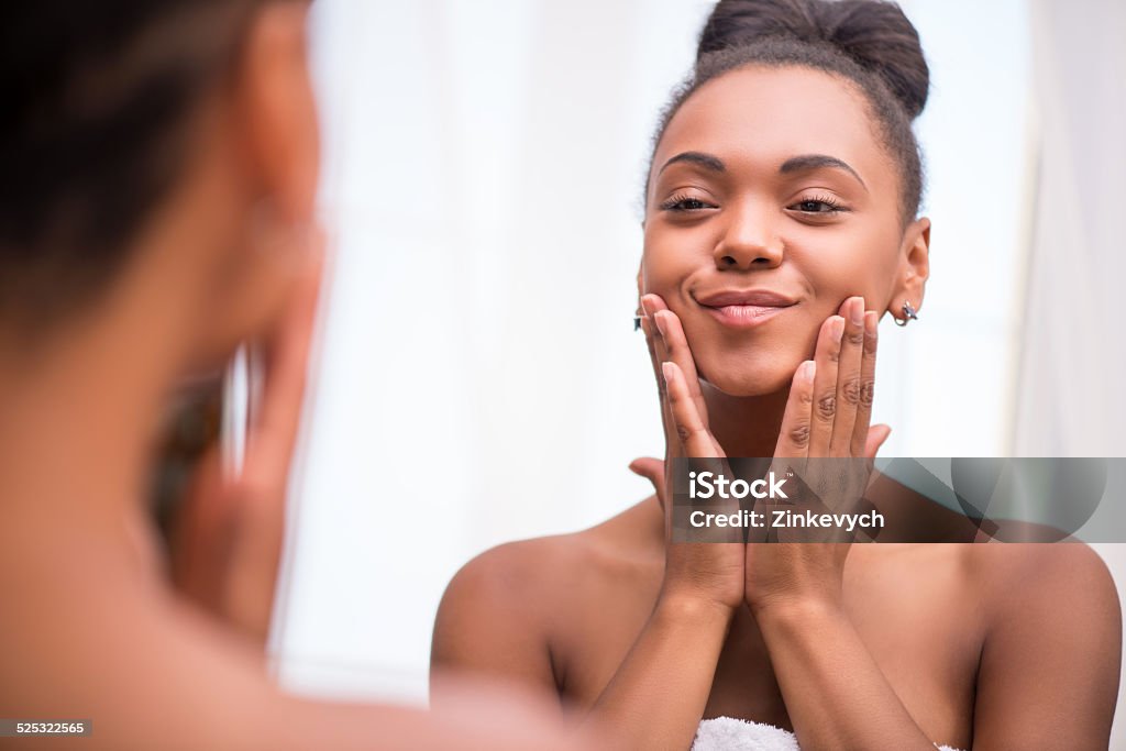 Beautiful  dark skinned girl in white towel Beautiful  dark skinned girl in white towel bringing face cream looking at mirror admiring  herself  touching cheeks smiling  isolated on white background Adult Stock Photo