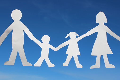 Photo showing a family paper chain of people (fathern, son, daughter and mother), cut out of white paper.  The people paper chain is pictured being held  in the strong afternoon sunshine (without the hands and fingers being visible in the photograph), isolated against a rich blue sky, free of clouds.  This is a concept photo for happy families - parents and their young children - adult man and woman, young boy and girl / mixed brother and sister siblings.