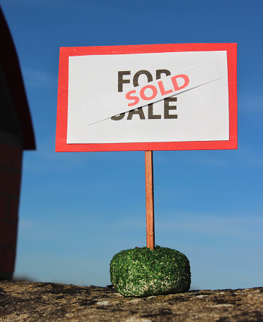 Photo showing a homemade model dolls house made from cardboard and coloured paper.  Standing outside of the front door is a For Sale sign - black writing on a white background, with a red 'Sold' banner.   The toy cardboard house and For Sale / Sold sign are pictured reflecting in the mirror, with a rich blue sky background (rather like it is reflecting in a pond, against the water's surface.