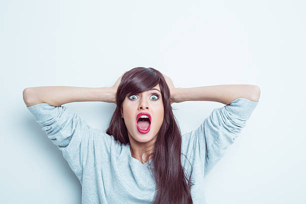 Shocked young woman Portrait of shocked young woman wearing grey jumper, raising her arms and shouting at camera, rolling eyes. Studio shot, white background. rolling eyes stock pictures, royalty-free photos & images