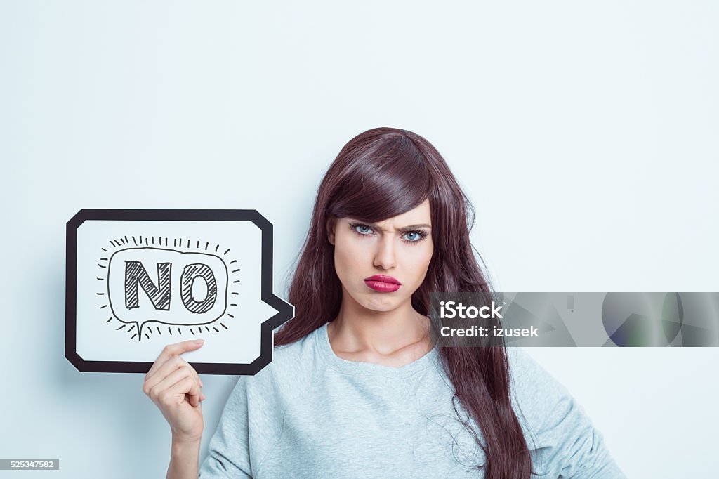 Unhappy young woman holding speech bubble Portrait of young woman wearing grey jumper, holding a speech bubble with word "no". Studio shot, white background. Single Word No Stock Photo