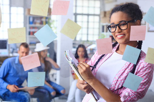 Cheerful young woman with eyeglasses and braids writing notes on papers with her left hand, standing behind a glass wall with colored sticky notes, in front of business team brainstorming. Focus on foreground.