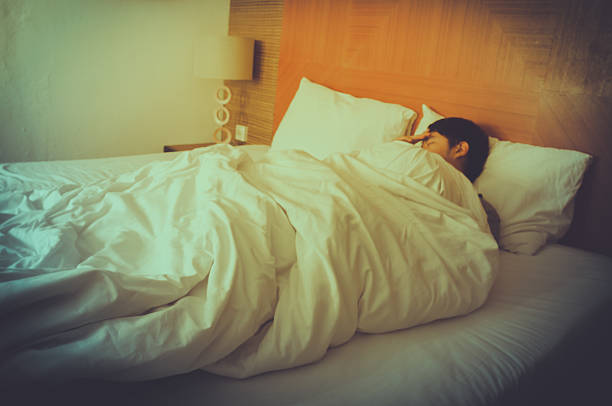 Stock photo showing Indian man wearing white towelling robe stood stretching in front of hotel window with open curtains looking out of window with a close-up view of retro, double bell, analogue alarm clock sitting on bedside table.