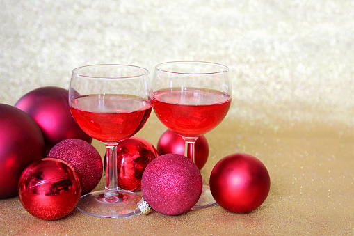 Two wine glasses fill with red beverage are sitting in a toast surrounded by Christmas tree bulb decorations in front of a glittery gold background.