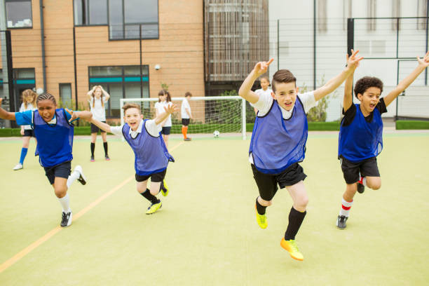 Schoolboys wearing sport uniforms running with arms raised in soccer field in front of school  recess soccer stock pictures, royalty-free photos & images