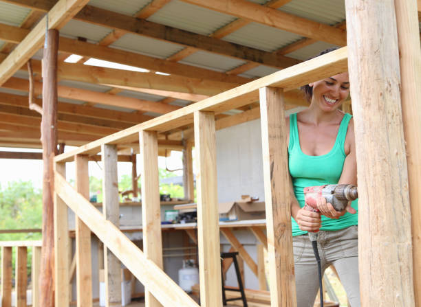 istock Smiling woman drilling in plank in house under construction 525440585