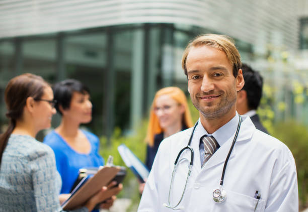 porträt eines lächelnden arztes im laborkittel vor dem krankenhaus, frauen mit klemmbrettern im hintergrund - healthcare and medicine smiling group of people lab coat stock-fotos und bilder