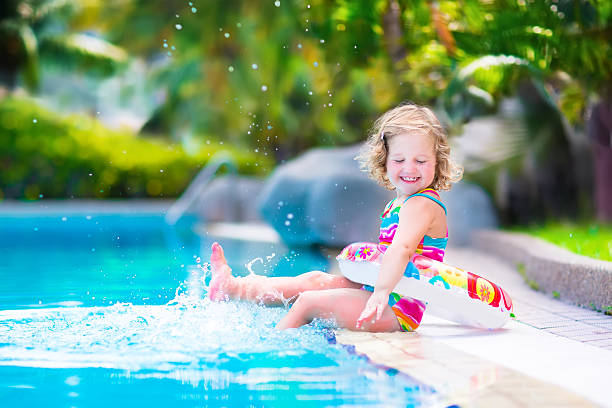 Little happy girl in a swimming pool Adorable little girl with curly hair wearing a colorful swimming suit playing with water splashes at beautiful pool in a tropical resort having fun during family summer vacation one piece swimsuit photos stock pictures, royalty-free photos & images