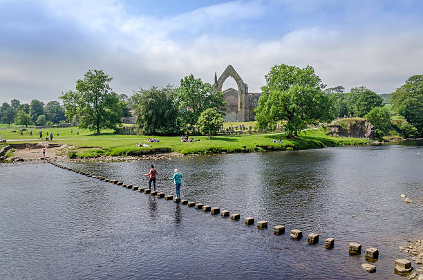 Bolton Abbey Stepping Stones Skipton, UK - July 18th, 2014; Tourists on the stepping stones across the River Wharfe next to the ruins of Bolton Abbey in the Yorkshire Dales wharfe river photos stock pictures, royalty-free photos & images