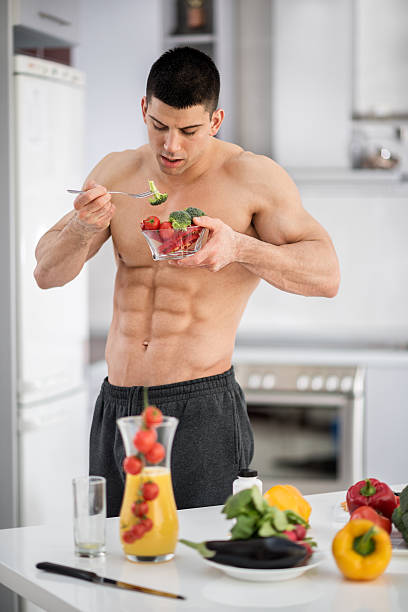 Shirtless athlete in the kitchen Young shirtless male fitness athlete standing in the kitchen and eating raw vegetables. There are kitchen knives, orange juice and more vegetables on the table in front of him. eating body building muscular build vegetable stock pictures, royalty-free photos & images