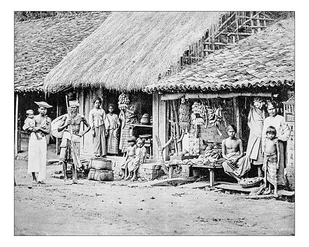 Antique photograph of group of native people from Sri Lanka during the 19th century, when it was called Island of Ceylon and it was a Bristish colony. A group of people of all ages, from baby to old, pose for a picture in front of their shacks and shops  