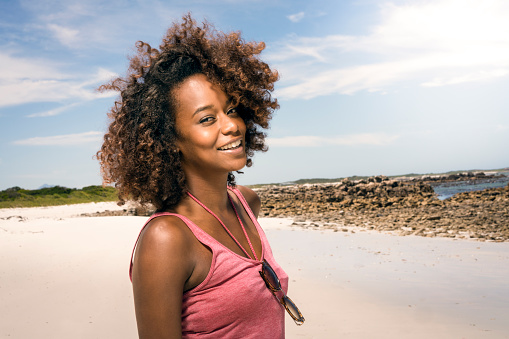 A photo of young woman smiling at beach. Portrait of confident attractive female is enjoying vacation. She is wearing tank top.