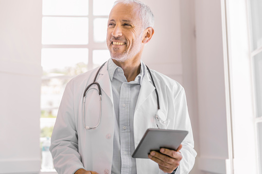 A photo of happy male doctor holding digital tablet in clinic. Thoughtful medical professional is wearing lab coat. Mature healthcare practitioner standing at brightly lit office.