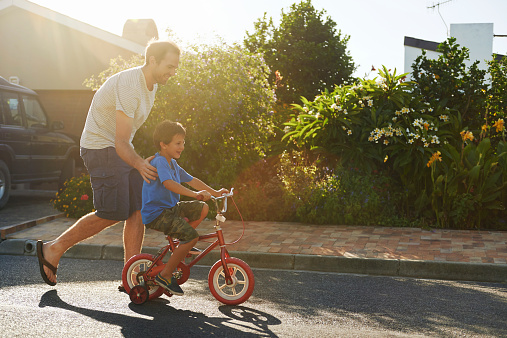 young boy learning to ride bicycle as father teaches him in the suburb street having fun.