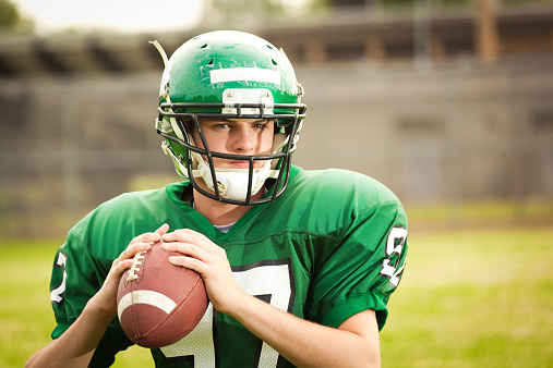 A high school quarterback American football player, gripping a ball, ready to throw a pass. The teenager boy wears a helmet and green padded uniform, with an outdoors stadium in soft focus in the background. The  young man aspires to play on a Midwest university or college competitive sports team.