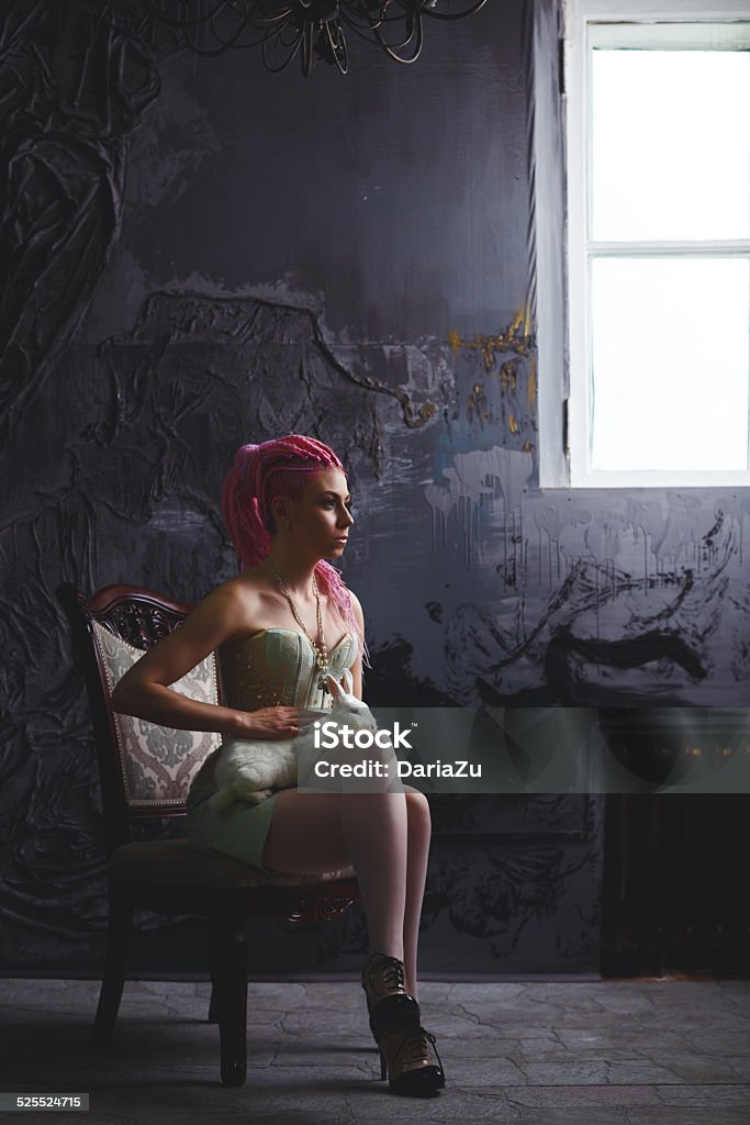 Young woman with dreadlocks wearing corset Freaky luxury woman in vintage corset sitting on ancient chair near window with white rabbit on her knees. Bright pink dreadlocks hairstyle, beauty make up. Gothic creative style. Adult Stock Photo