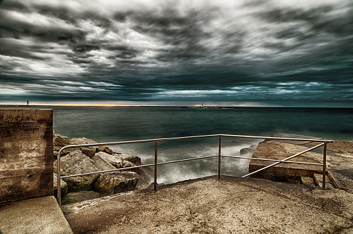Sunset on breakwater with stairs on the Mediterranean sea with red steel lookout and green top white lighthouse in the cloudy background near Porec in Croatia