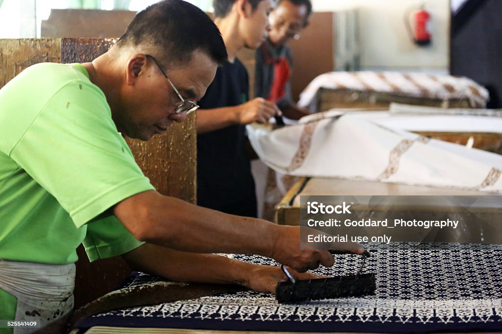 Indonesia: Batik Making in Yogyakarta Yogyakarta, Indonesia - August 14, 2014: Two men use a copper stamp called a cap to apply wax in patterns onto a cloth to make a batik. Batik is a traditional form of decorating cloth, and Java is particularly famous for its variety of patterns and the quality of workmanship. The technique includes the manual application of wax between each dye bath to add each layer of colour individually. Art Stock Photo