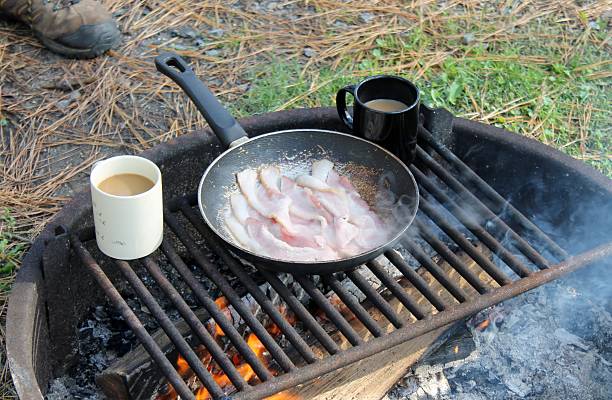 Warming cups of coffee on the camp fire while the bacon cooks in a skillet. Fire ring with a grate holding fire wood and utensils as smoke rises off to the side. Black and white mugs and worn skillet.  Pine needles carpet ground.