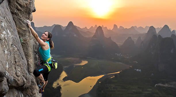 Female climber on Chinese background Female extreme climber conquers steep rock against the sunset over the river. China, typical Chinese landscape with mountains and river rock climbing stock pictures, royalty-free photos & images