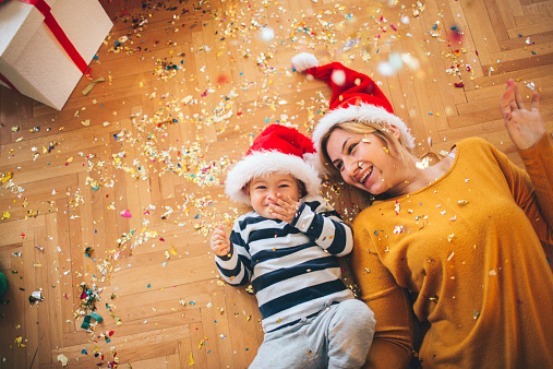 Little boy decorating his first Christmas tree, along with his mother, puting an ornaments and lights, playing along with conffeti an laughting merrily