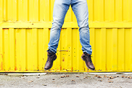 Male legs in the middle of jump in blue jeans and brown leather boots in front of bright yellow metal door. Urban scene.