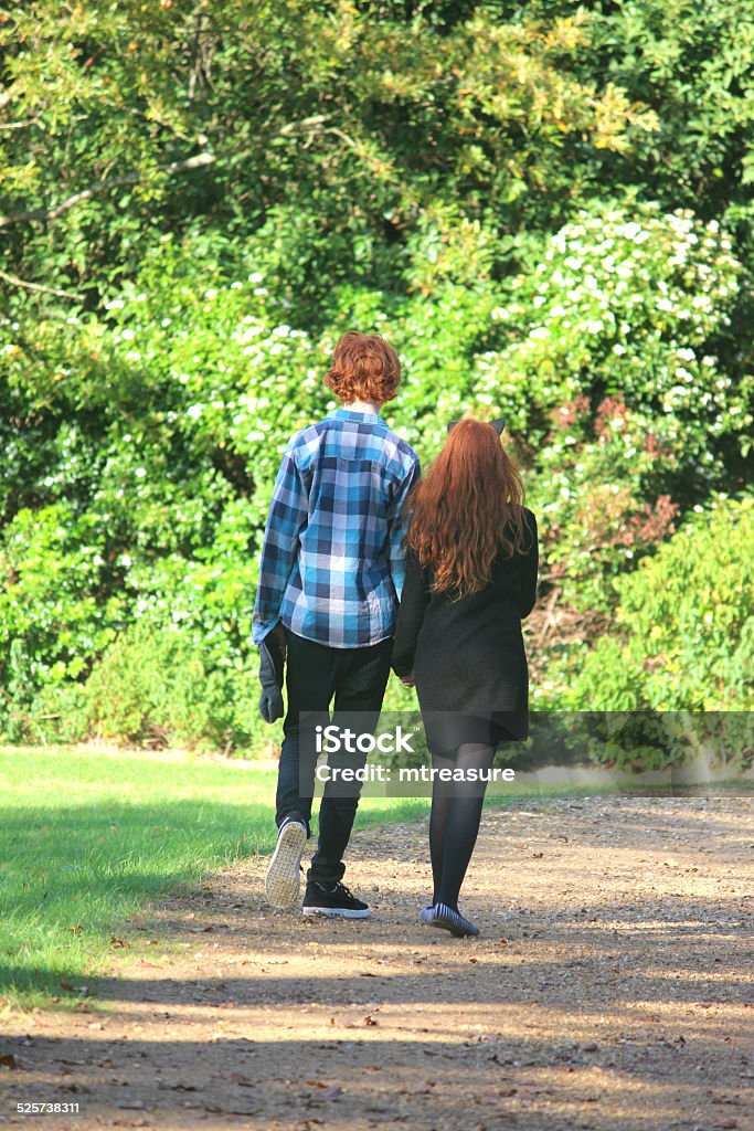 Boy and girl walking along woodland pathway / footpath, holding-hands, red-hair Photo showing a teenage boy and girl walking along woodland pathway / footpath, holding hands.  The boy and girl are actually brother and sister (siblings), messing around on a sunny afternoon.  They both have hair ginger / red hair and are pictured in the sunshine, with shadows from the surrounding trees falling on the path and lawn / grass. Beauty In Nature Stock Photo