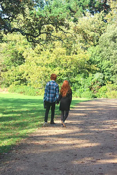 Photo showing a teenage boy and girl walking along woodland pathway / footpath, holding hands.  The boy and girl are actually brother and sister (siblings), messing around on a sunny afternoon.  They both have hair ginger / red hair and are pictured in the sunshine, with shadows from the surrounding trees falling on the path and lawn / grass.