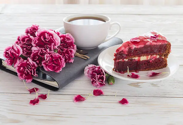 Red velvet cake, cup of coffee, notebook and pink carnations on wooden table