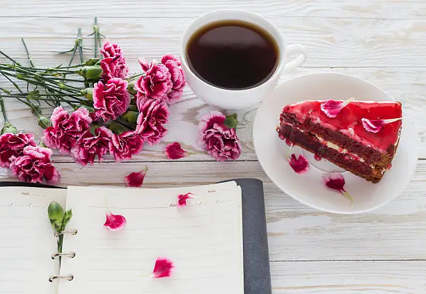 Red velvet cake, cup of coffee, notebook and pink carnations on wooden table