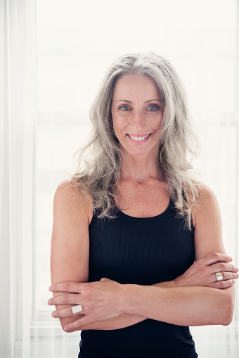 Toned and tanned mature woman with silver hair in tanktop. She is standing in front of a window, arms crossed, looking at camera with a smile. Sporty look with natural makeup. Vertical backlit indoors waist up shot, with copy space. This was taken in Quebec, Canada.