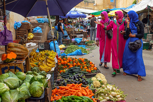 Fes, Morocco - April 10th, 2014: Three Muslim women in pink and blue traditional clothes looking at fruit and vegetable at Old Medina market in the city of Fes, Local people in the background selling and buying goods. Morocco, North Africa.