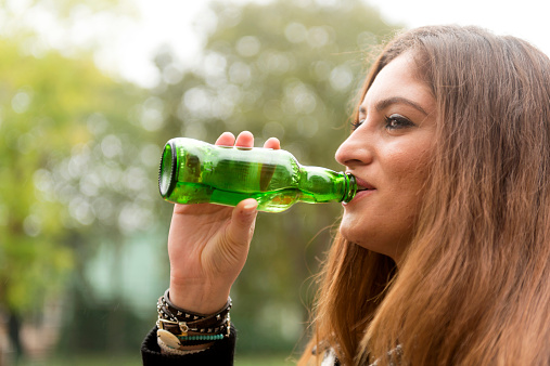 Beautiful Turkish student with long brown hair drinking soft drink from a green bottle during break at secondary school, university,  Istanbul, Turkey, outdoors, day, drizzling. Nikon D800, full frame, XXXL.