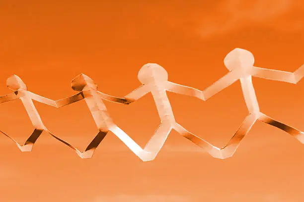 Photo showing a people paper chain of dolls, showing a row of white paper men holding hands.  The paper chain is pictured being held up in mid air, isolated against an orange background.  This is a concept photo to cover a variety of themes, such as teamwork, unity, relationships, harmony, friendship and general togetherness.