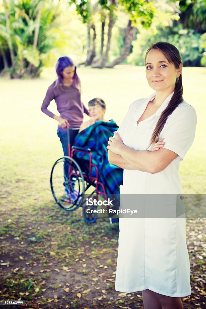 Smiling nurse enjoys outing for elderly patient and young friend A smiling, confident young nurse stands in front of her patient - a wheelchair-bound old woman - and a young relative, as they all spend some quality time in a sunny park. She really enjoys her work. 20-29 Years Stock Photo
