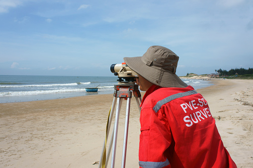 Binh Thuan, Viet Nam - October 21, 2014: Asian engineer work on Vietnamese beach, man looking in theodolite to survey sea level, measurement device set on tripod, Vietnam, Oct 21, 2013