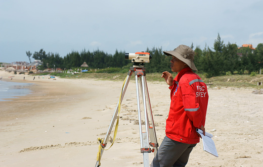 Binh Thuan, Viet Nam- October 21, 2014: Asian engineer work on Vietnamese beach, man looking in theodolite to survey sea level, measurement device set on tripod, Vietnam, Oct 21, 2013