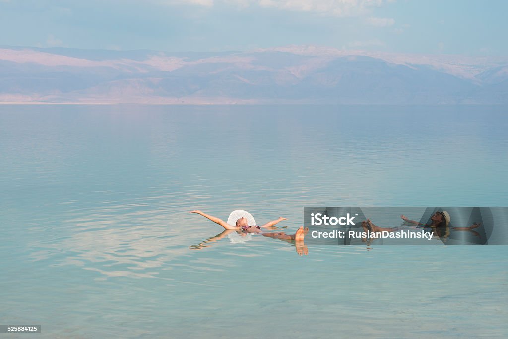 Vacation in Dead Sea with friends. Lovely multiracial girls wearing sun hats, lying on back with outstretched arms, floating in salty water of Dead Sea. Unusual buoyancy caused by high salinity. Dead Sea Stock Photo