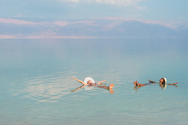 Vacation in Dead Sea with friends. Lovely multiracial girls wearing sun hats, lying on back with outstretched arms, floating in salty water of Dead Sea. Unusual buoyancy caused by high salinity. historical palestine photos stock pictures, royalty-free photos & images