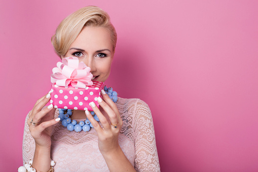 Happy birthday. Sweet blonde woman holding small gift box with ribbon. Soft colors. Studio portrait over pink background