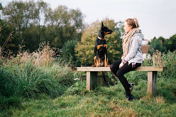 blonde girl and black dog sitting on bench facing each other.