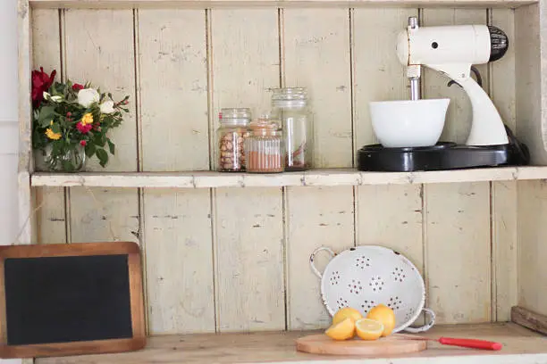 Arrangement of a few kitchen items on an antique farmhouse kitchen hutch made of distressed wood. There are some roses from the garden, spices, an old fashioned mixer, blackboard and a colander with lemons.