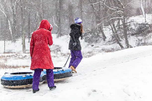 Two sisters have fun with snowtubing at the winter resort. They go to a hill top, oldest sister carry the tube. 